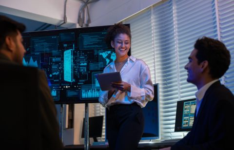 Group of professionals businesswoman and businessman in formal attire discussing ideas on innovative technology development project with tablets and laptops in dark modern office